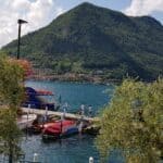 Lago d'Iseo: vista panoramica di Sulzano con Monte Isola sullo sfondo, barche ormeggiate e ulivi in primo piano.