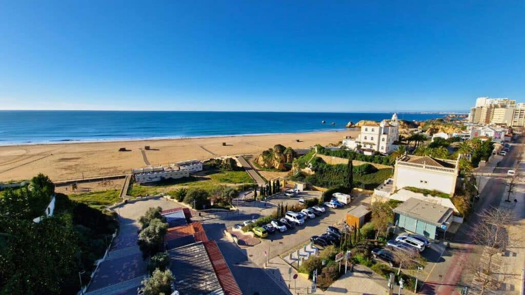 Come spostarsi in Algarve? Noleggiare l'auto dà una svolta alla tua vacanza! 2 Spiaggia dorata di Praia da Rocha in Algarve con edifici bianchi e auto parcheggiate, suggerendo la comodità di un'auto a noleggio per esplorare.