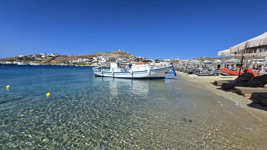 Spiaggia di Ornos Mykonos: barca ancorata nell'acqua cristallina, con i lettini posti così a riva che le onde arrivano quasi a toccarli.