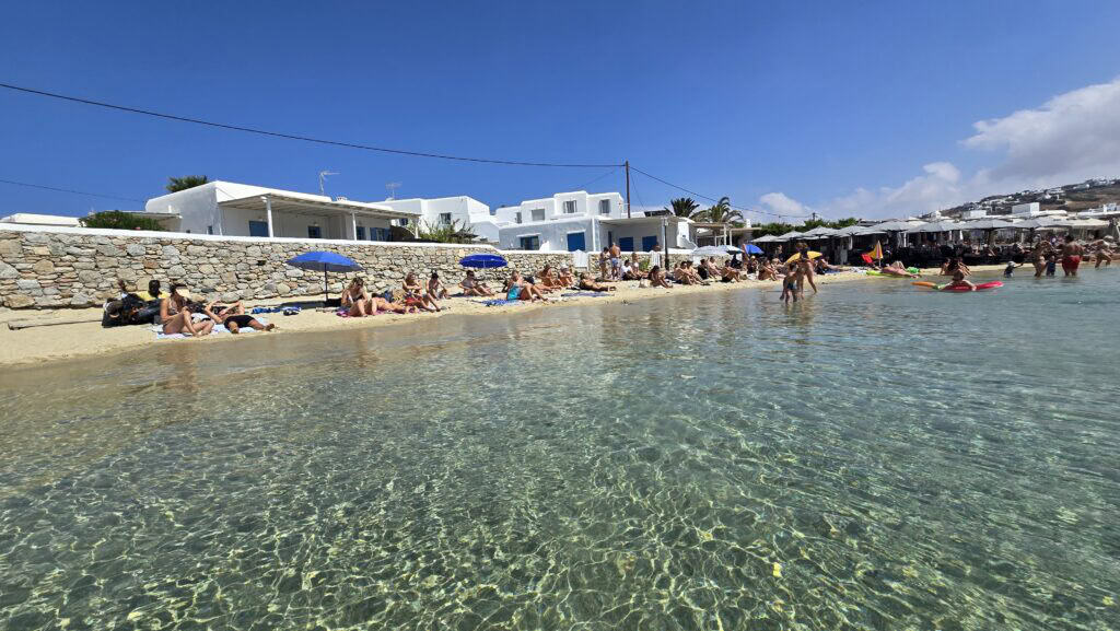 Area limitata di spiaggia libera a Ornos Beach Mykonos, con acqua cristallina e bagnanti che prendono il sole vicino alle case bianche dell'isola.