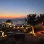 Vista mozzafiato del tramonto di Mykonos da un bar in collina, con una scultura a forma di cuore illuminato e un classico mulino a vento.