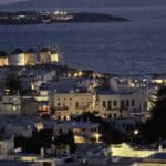 Vista panoramica di Mykonos di notte, con gli iconici mulini a vento illuminati sulla collina che domina il mare.