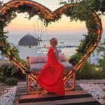 Una donna in abito rosso in posa davanti a un'installazione luminosa a forma di cuore in un sunset bar di Mykonos con il mare e un mulino a vento sullo sfondo.