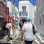 Persone che camminano sulle famose strade di ciottoli bianchi e grigi di Mykonos Town, con negozi e balconi rossi sullo sfondo.