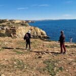 Ila e Gabriel di Tripilare (Gabriel è quello con i pantaloni rossi) vestiti con pantaloni lunghi e giubbini durante una passeggiata mattutina su un sentiero costiero roccioso in Algarve a Gennaio, con vista sull'oceano.