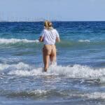 Turista con maglietta e sciarpa in costume da bagno entra nell'acqua fredda dell'Oceano Atlantico a Playa de Maspalomas, Gran Canaria, in pieno inverno (gennaio).