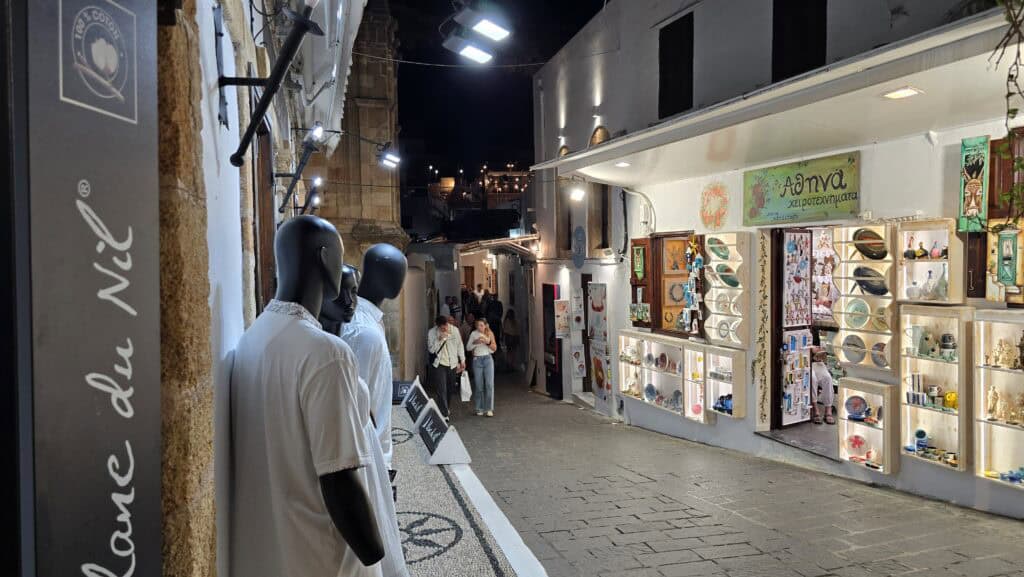 🌅 Lindos di Sera: Tra Acropoli Illuminata e Cene Romantiche sui Tetti di Rodi 3 Via principale di Lindos (Rodi) di sera, illuminata, con persone che passeggiano e boutique aperte. Illustra l'atmosfera vivace del centro storico.