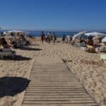 Passerella di legno sulla spiaggia di Praia da Rocha a Portimão in Algarve ad agosto. La spiaggia è affollata con file di lettini e ombrelloni bianchi, evidenziando il forte contrasto con l'inverno.