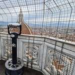View of Florence city from the top of Giotto's Bell Tower, showing the panoramic cityscape as seen through the protective security mesh/net. A coin-operated telescope is also visible on the viewing platform.
