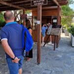 Gabriel walking towards the ticket area with the 'Tour Guide' sign visible above the entrance at Knossos Palace.