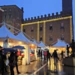 Night view of the Torrone Festival in Cremona with illuminated historic buildings and white stalls, showing visitors with umbrellas walking in the rain.