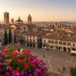 Scenic view of Piazza Vecchia in Bergamo's Città Alta, Italy, featuring historic architecture, Duomo dome, bell towers, and colorful flowers at sunset.