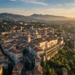 Panoramic aerial view of Bergamo's Città Alta, Italy, featuring historic buildings, the Duomo's dome, terracotta roofs, and surrounding hills at sunrise.