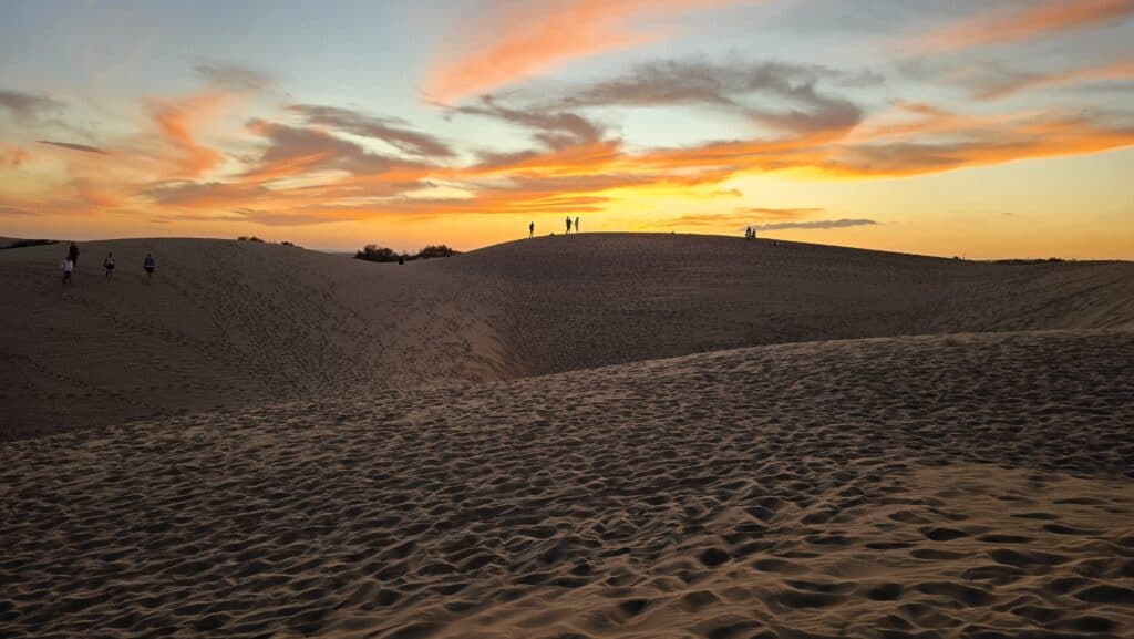 Hikers silhouetted against a fiery sunset sky while walking along the crests of the Maspalomas Dunes in Gran Canaria, Spain.