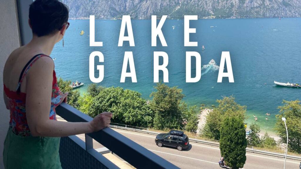 Woman on a balcony overlooking Lake Garda with a car driving along the road below and mountains in the background.