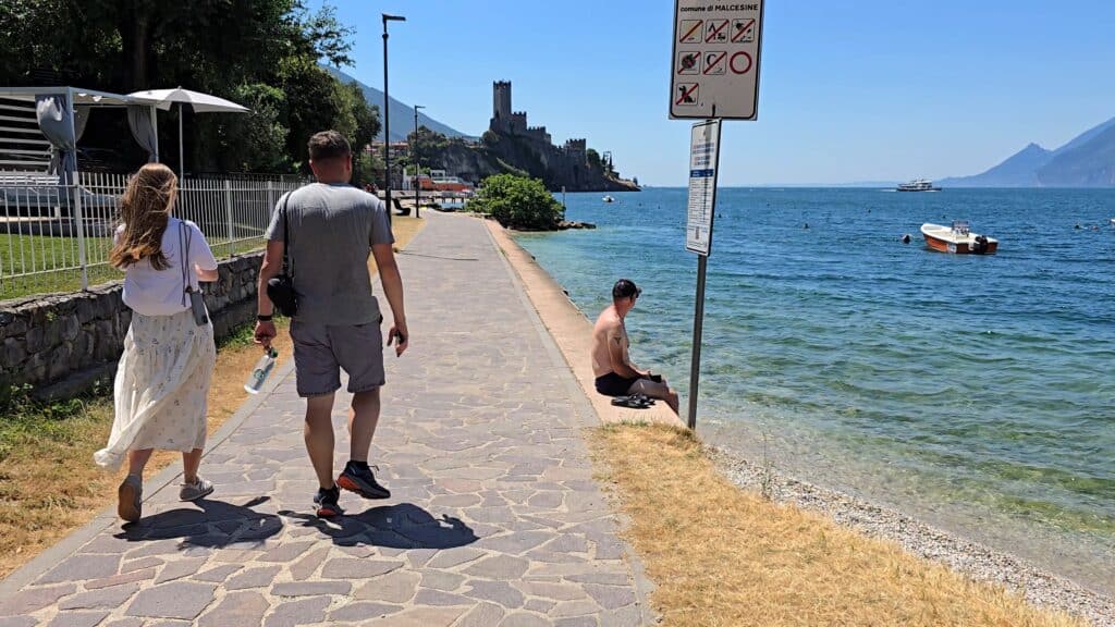 Lake Garda Hikes: Your Ultimate Guide to Breathtaking Trails & Scenic Walks 1 **Lake Garda Hikes**: People walking on a paved promenade next to the clear blue waters in Malcesine, with the Scaliger Castle and distant mountains in the background.