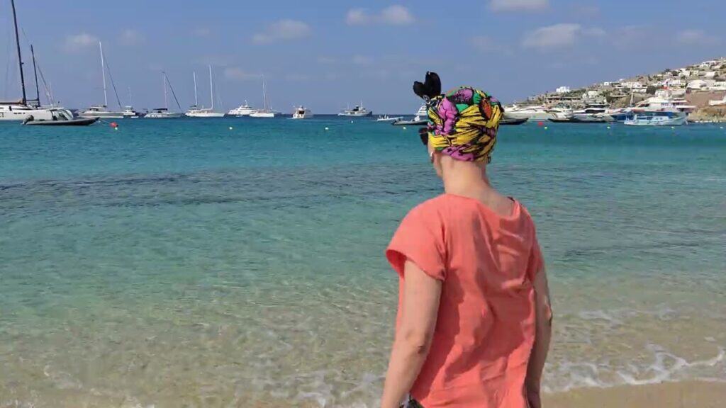 A person standing on Platis Gialos beach in Mykonos, looking out at the sea and boats anchored in the distance.