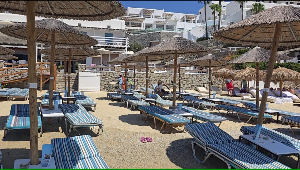 Cavo Psarou sunbeds and umbrellas on Psarou Beach, Mykonos, showing the beach club setup and the white Cavo Psarou Suites accommodation in the background.