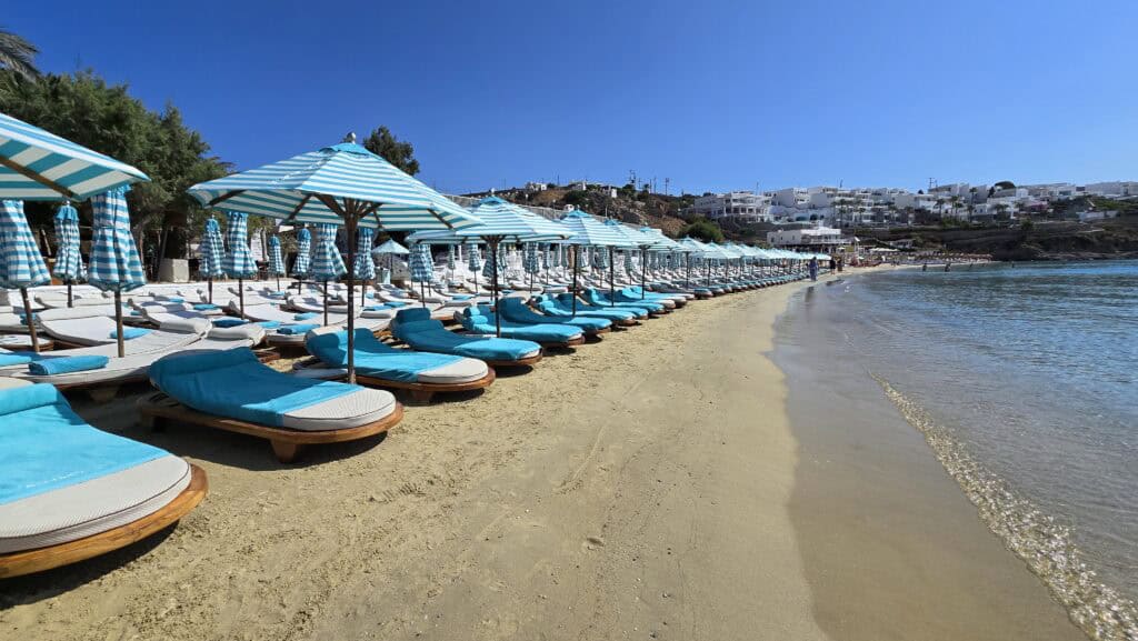 View of the front row of luxury sunbeds at Nammos Beach Club, Psarou Mykonos, with striped umbrellas and crystal-clear sea.