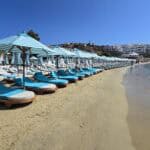 View of the front row of luxury sunbeds at Nammos Beach Club, Psarou Mykonos, with striped umbrellas and crystal-clear sea.