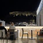 Couple enjoying the CourtYard rooftop By Night with a panoramic view of the illuminated Acropolis. Shows elegant and smart-casual dress code mix.