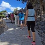 A group of tourists, including Gabriel (wearing a blue backpack), walk uphill on a wide, paved stone path towards the Acropolis of Lindos, with comfortable shoes visible.