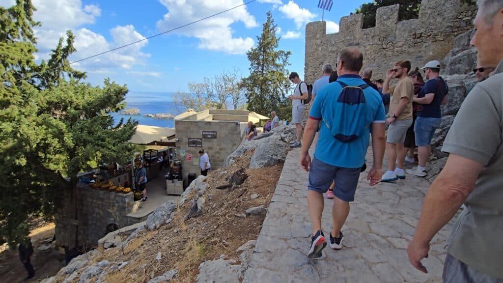 Tourists, including Gabriel with a blue backpack, walk past a bar with outdoor seating and a scenic view of the sea and Lindos village below, just before the Acropolis entrance.