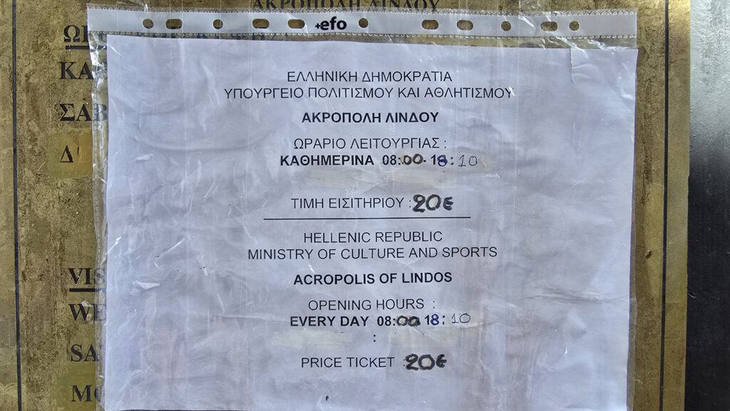Official sign from the Hellenic Ministry of Culture for the Acropolis of Lindos, showing opening hours (08:00–18:10) and the ticket price (20€), with handwritten corrections.
