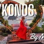 An elegant woman in a red dress poses in front of a heart-shaped light installation and a windmill at sunset, showing the charming side of Mykonos.