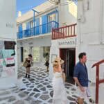 People walking on the famous white and gray cobblestone streets of Mykonos Town, with shops and blue balconies in the background.