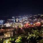A wide panoramic night view of Mykonos, showing the illuminated Little Venice waterfront, the windmills, and cruise ships in the harbor.