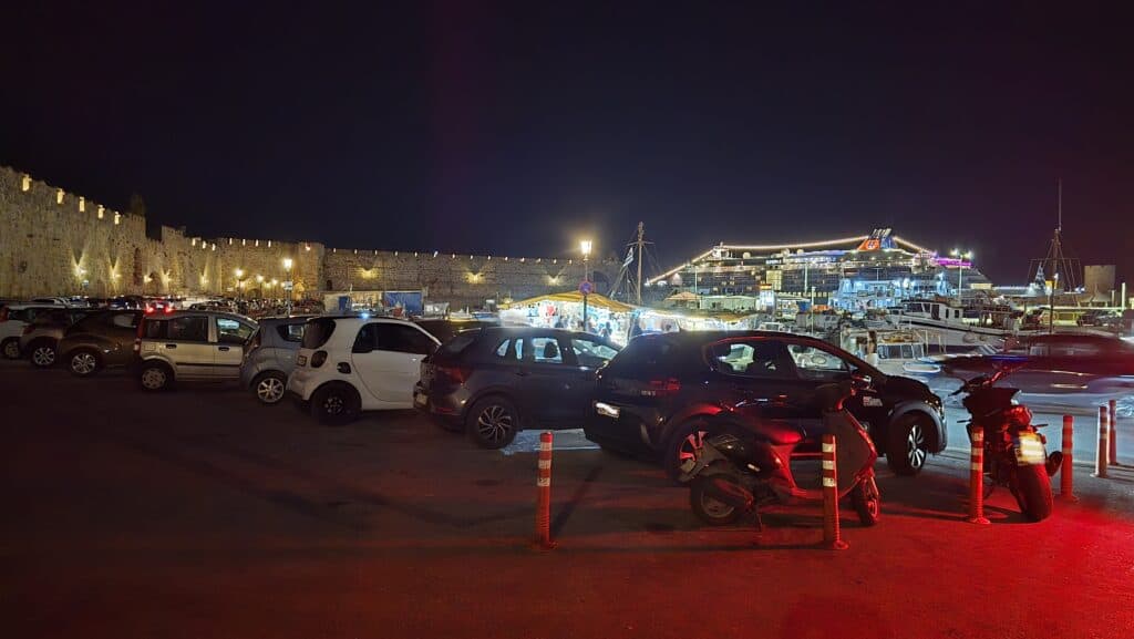 Night view of the Rhodes Old Town medieval walls and a crowded parking lot next to a brightly illuminated cruise ship at the port.
