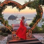 A woman in a red dress posing in front of a heart-shaped light installation at a Mykonos sunset bar with the sea and a windmill in the background.