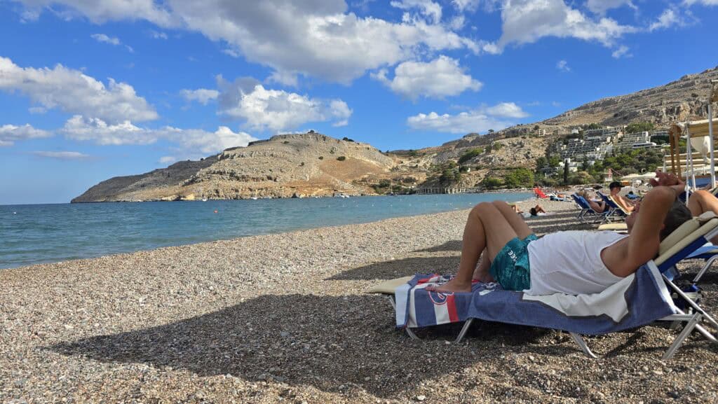 A man relaxing on a sunbed on the pebble beach of Vlicha Bay, Rhodes, with the calm sea and surrounding hills visible under a blue sky with clouds.