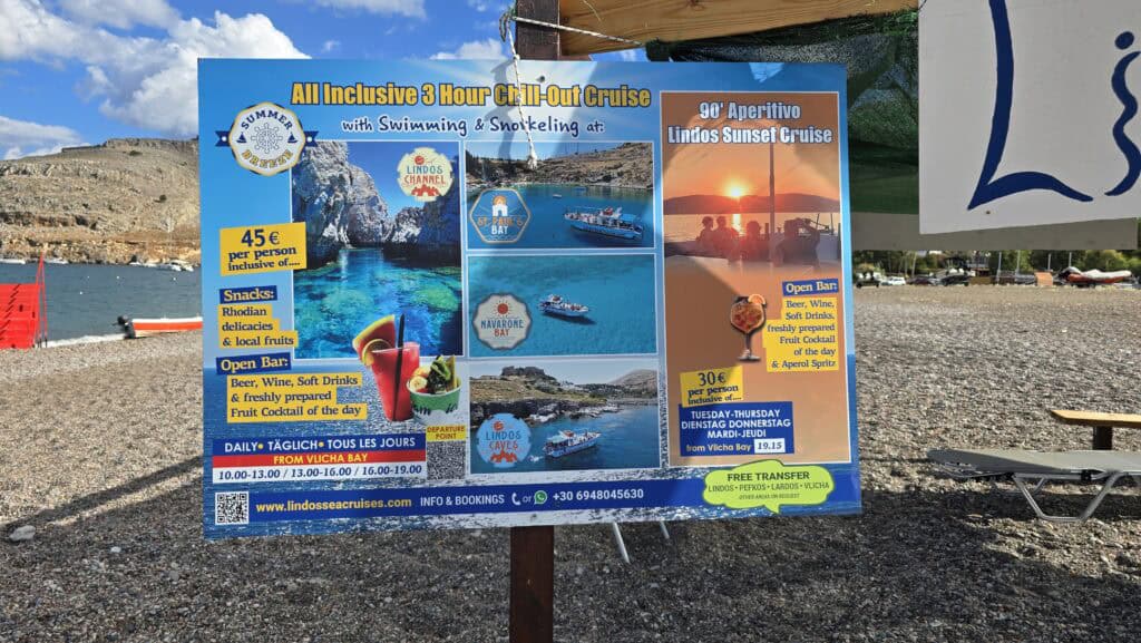 A seaside billboard for Lindos Sea Cruises detailing excursions from Vlicha Bay, including the 3-hour Chill-Out Cruise and the Lindos Sunset Cruise, with prices of €45 and €30 respectively for October 2025.