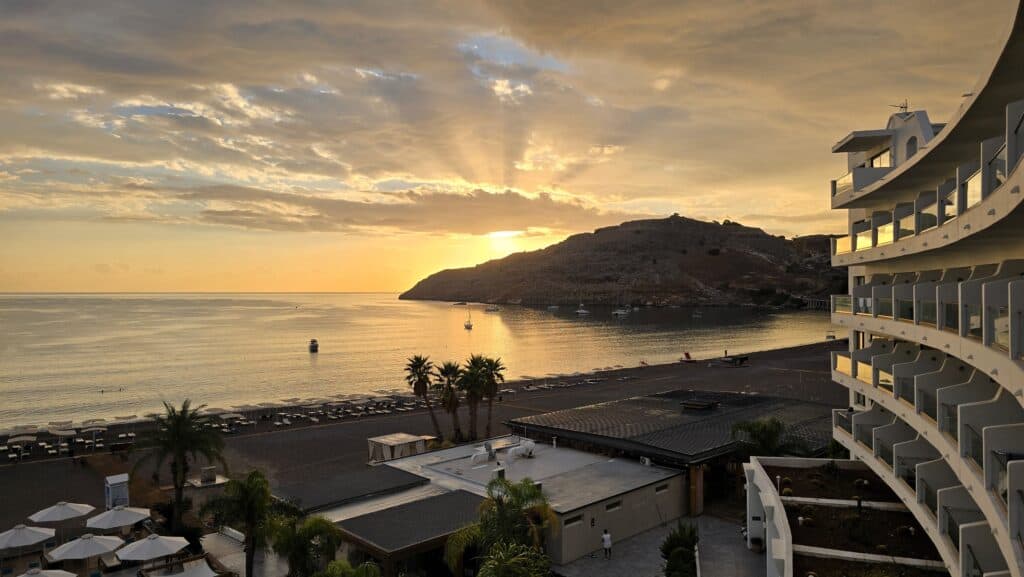 Golden sunrise over Vlicha Bay, Rhodes, seen from a balcony of the Lindos Bay Hotel, with the sun cresting behind the headland and casting rays over the calm water and the deserted beach below.