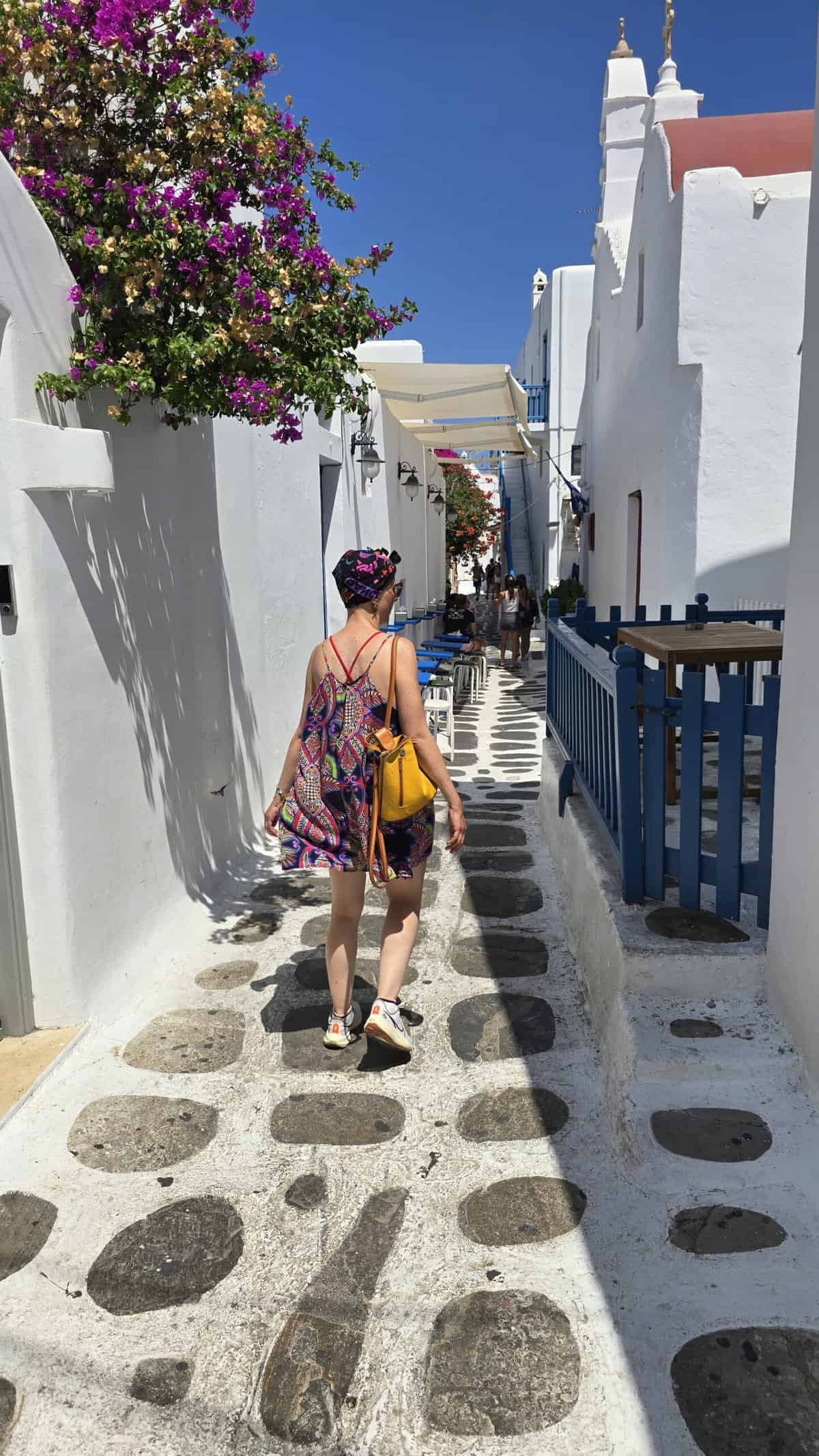 Mykonos Town (Chora) Unfiltered: A Guide to Sunsets, Windy Adventures, and Hidden Corners 5 The author, Ila, seen from behind as she walks through a narrow, charming alley in Mykonos Town, featuring traditional white houses, blue wooden railings, and vibrant pink bougainvillea flowers.