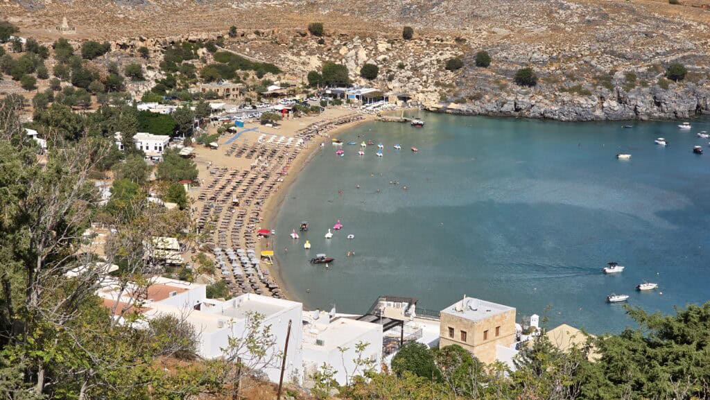 An aerial view of Lindos Main Beach (Megali Paralia), perfectly illustrating its expansive crescent shape, the blend of organized sunbeds, and free beach space available for visitors.