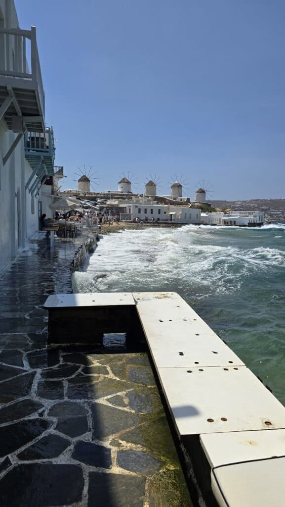 Mykonos Town (Chora) Unfiltered: A Guide to Sunsets, Windy Adventures, and Hidden Corners 3 Occasional waves splashing onto the stone walkway of Little Venice in Mykonos Town, with the sea interacting with the waterfront cafes and the windmills in the background.