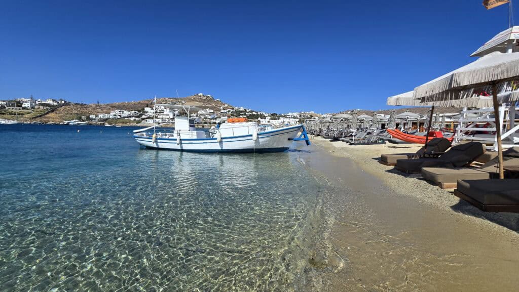 Stunning Ornos Beach, Mykonos, featuring a traditional Greek boat on crystalline water next to luxury sunbeds, highlighting the high quality despite limited walking space.