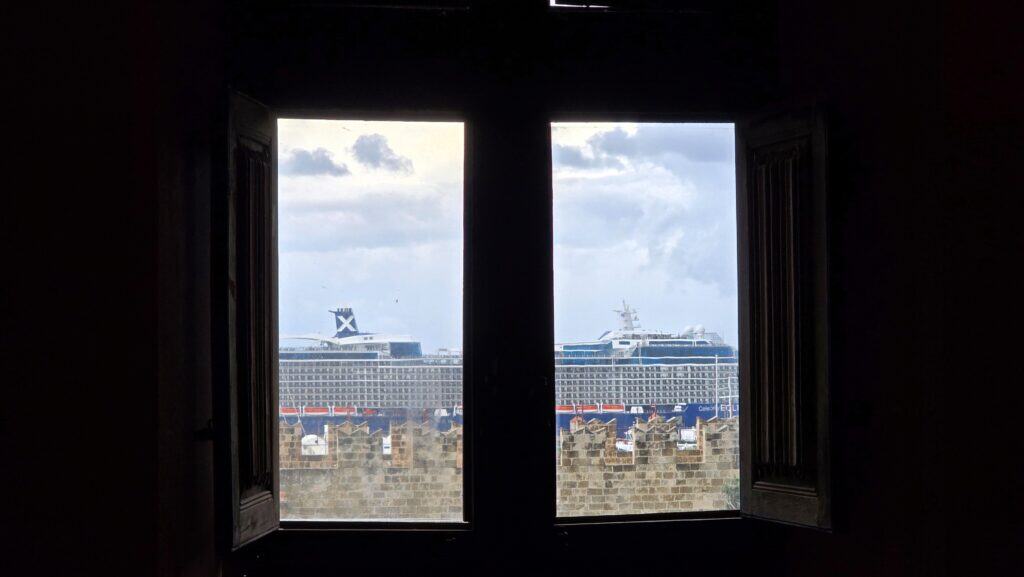View from a window of the Grand Master's Palace, where thick medieval walls frame a view of a modern cruise ship docked in the harbor of Rhodes City.