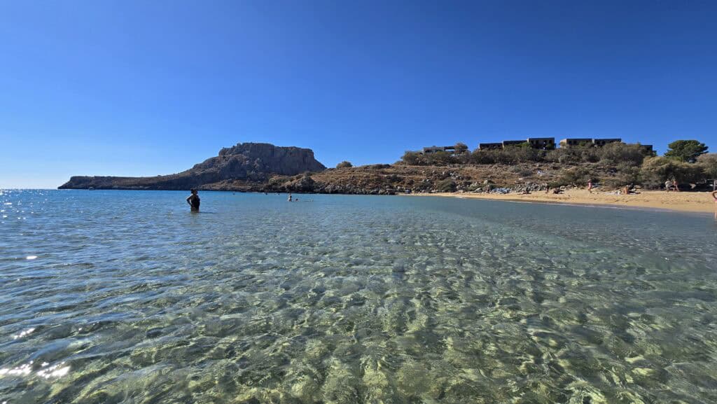 Agathi Beach Rhodes: The Essential Guide to the "Golden Sand" Gem 3 View of Agathi Beach highlighting the extremely shallow, crystal-clear water, with people standing far from the shore. This confirms Agathi as a safe, gently-sloping beach ideal for families and young children.