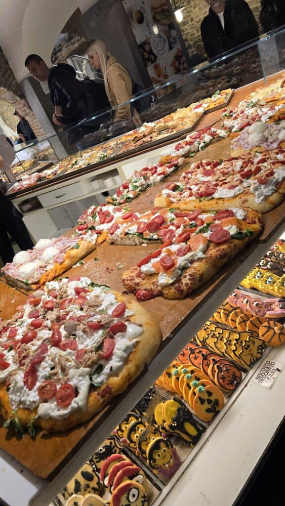 Street view of a vibrant window display in Bergamo Alta featuring slices of Italian pizza, focaccia, and seasonal decorated Halloween cookies priced at 2.50 Euros.