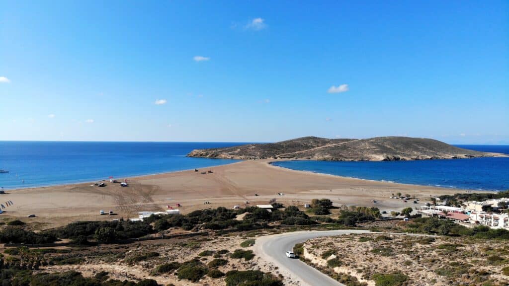 Aerial overview of Prasonisi, showing the curved road leading to the isthmus, the two seas meeting, and the free parking area, confirming the easy asphalt road access.