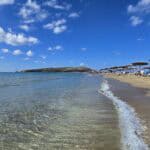 Calm, crystalline Mediterranean Sea side of Prasonisi, showing sunbeds and a small windsurf sail in the distance, illustrating the ideal conditions for beginners and relaxation.