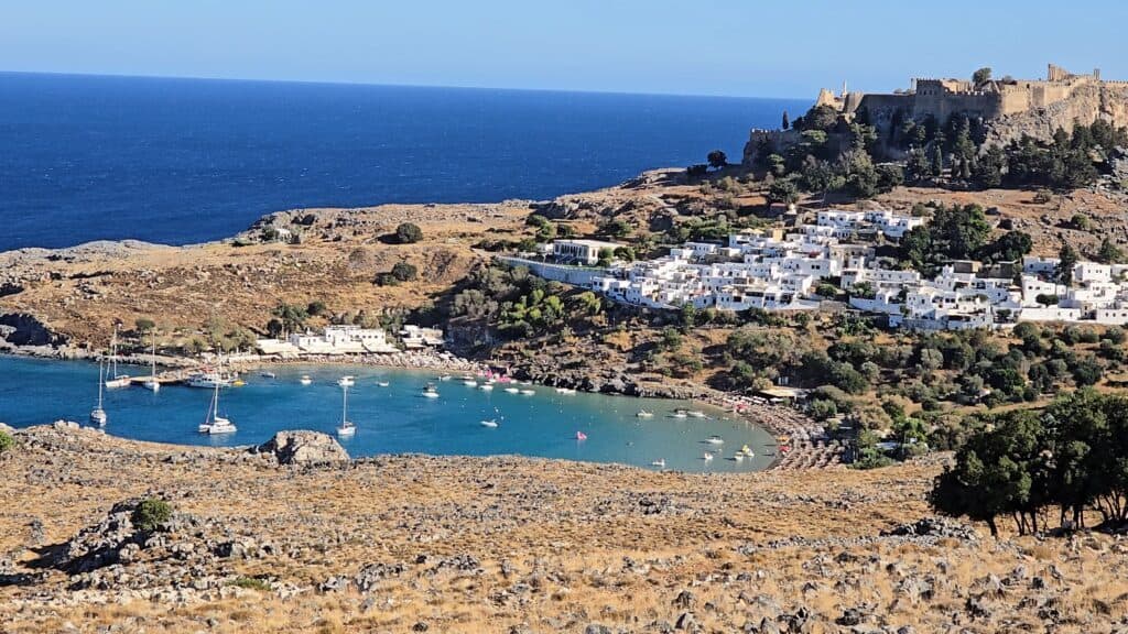 Panoramic view of Lindos Bay, Rhodes, showing both Lindos Main Beach and Pallas Beach with their clear blue waters, the whitewashed houses of Lindos village, and the ancient Acropolis perched on the hill above, all under a clear blue sky.