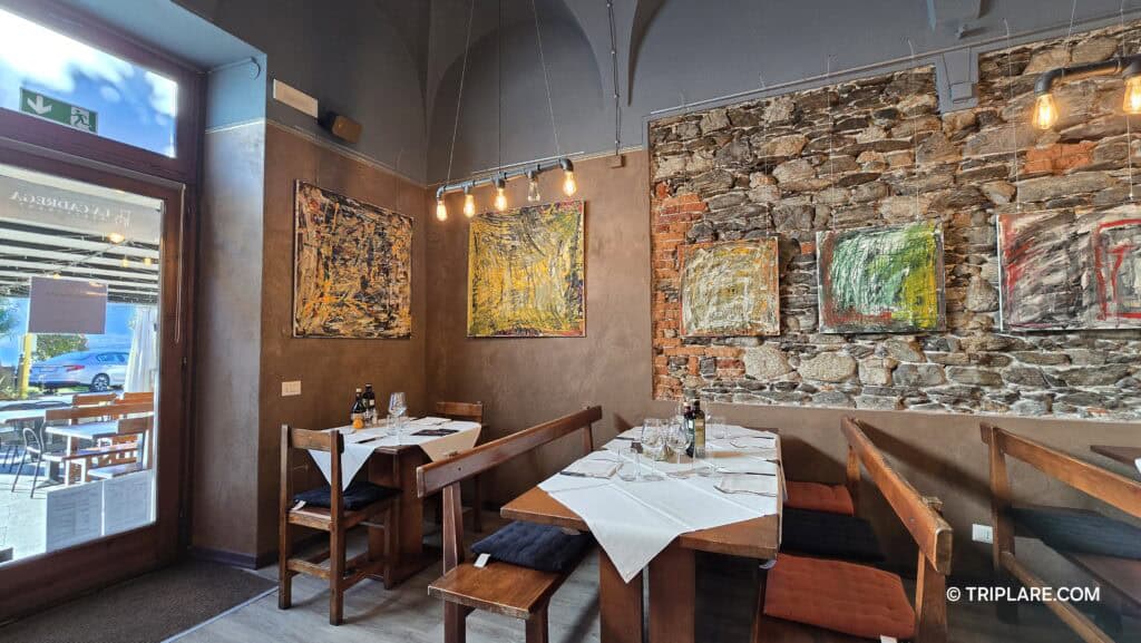 Interior of La Cadrega restaurant in Verbania Suna, showing rustic stone walls, contemporary art, and the street outside the window.
