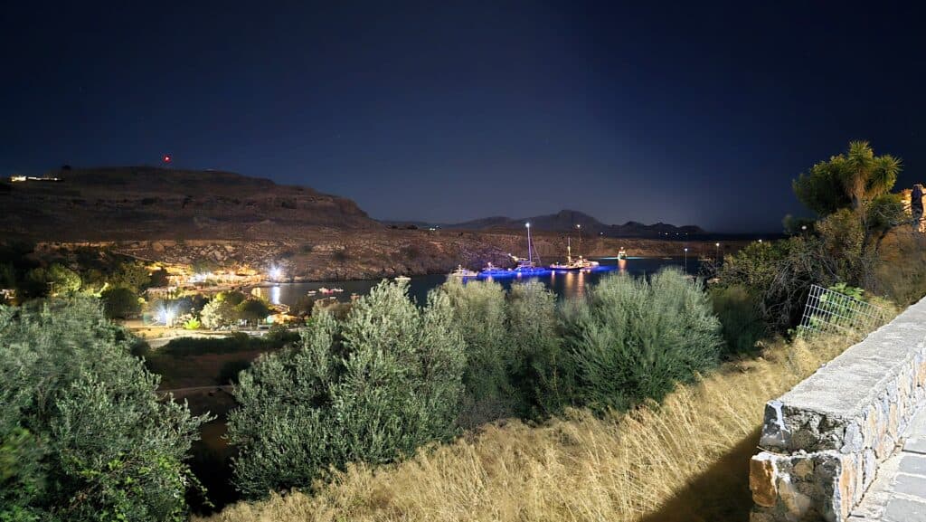 Panorama of the illuminated Lindos Beach (Paralia Lindos) area, viewed from the village center at night, showing the active shoreline where seaside restaurants are located.