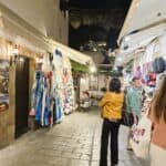 Illuminated narrow alleyway in Lindos at night, showcasing boutique shops with unique finds. The illuminated Acropolis is visible in the background, blending shopping with the iconic view.