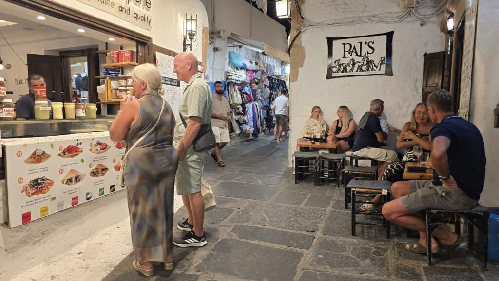 A lively evening scene on the pedestrian streets of Lindos, showing open shops, a street bar, and a place to buy evening treats like crêpes.